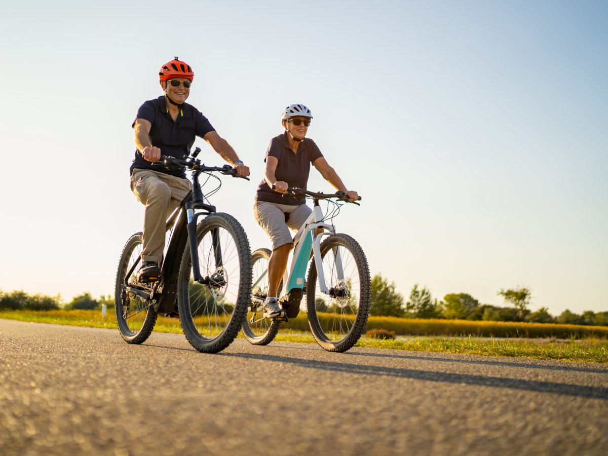 low perspective happy smiling fit senior couple cycling on electric bicycles on road through flat rural landscape on sunny summer day
